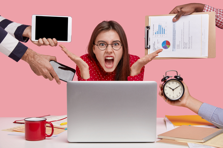 Annoyed freelancer gestures in front of laptop computer, has much paperwork, works overtime, cries desperately, drinks coffee, poses over pink background. Deadline and negative emotions concept
