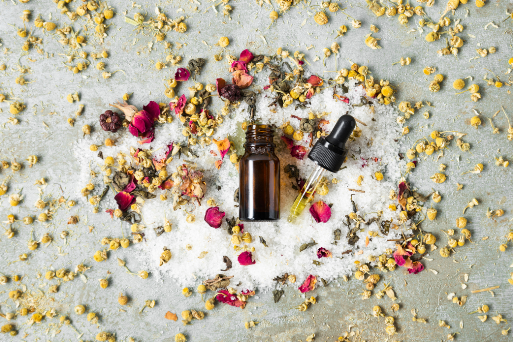 Vue de dessus d’une bouteille de remède naturel avec un compte-gouttes, entourée de fleurs séchées, de camomille et de pétales de rose sur un fond de sel de bain. Illustration des produits de bien-être naturels et des élixirs floraux, tels que les Fleurs de Bach, pour équilibrer les émotions.